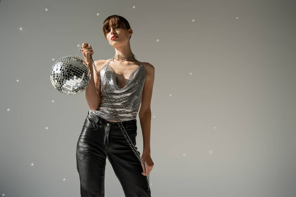 stylish woman in shiny top and leather pants holding disco ball on grey background  - Photo, Image