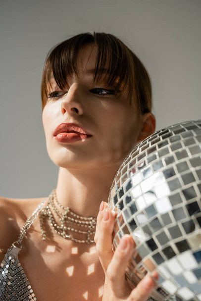 молодая женщина с челкой, держащая блестящий диско-шар изолирован на сером - Фото, изображение
