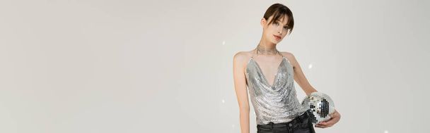 brunette young woman in trendy top and leather pants holding retro disco ball isolated on grey, banner   - 写真・画像