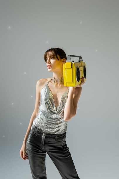 young woman in trendy top and leather pants holding yellow boombox on grey  - Foto, afbeelding