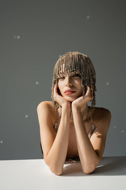woman with metallic headwear with jewelry looking at camera while sitting at desk on grey  - Foto, Bild