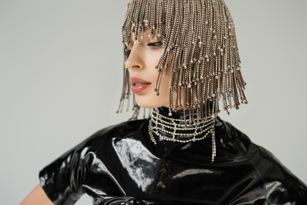 Fashionable woman in jewelry headwear and latex clothes looking away isolated on grey  - Foto, immagini