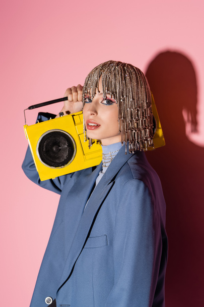 Trendy woman in jacket and jewelry headwear holding boombox on pink background  - Foto, Imagen