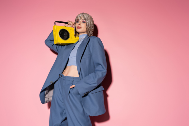 Stylish woman in suit and jewelry headwear holding boombox and posing on pink background  - Fotografie, Obrázek