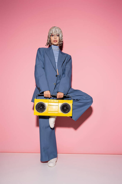 Full length of stylish model in jewelry headwear holding bright boombox on pink background  - Foto, imagen