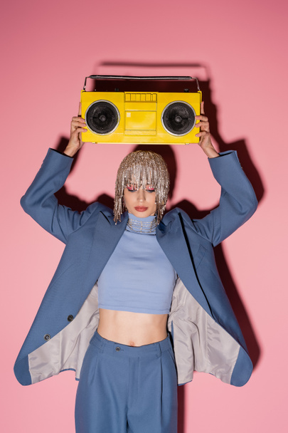 Fashionable woman in luxury headwear and suit holding boombox above head on pink background  - Foto, Bild