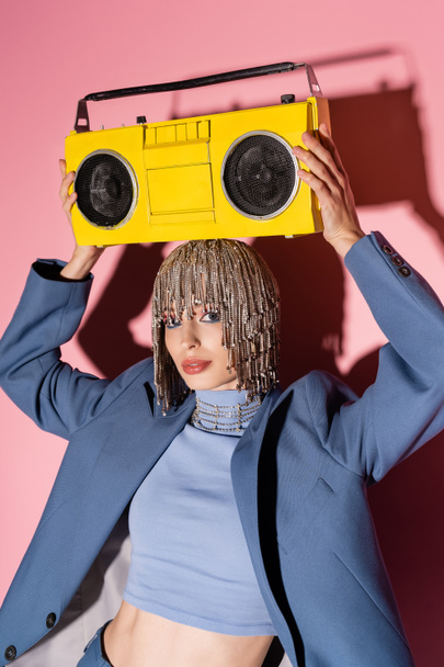 Stylish model in jewelry headwear and jacket holding boombox above head on pink background  - Fotó, kép