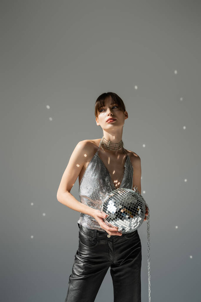 Модная молодая женщина в блестящем топе держит диско-мяч на сером фоне  - Фото, изображение