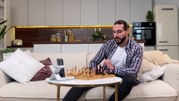 Middle adult man playing board game with friend via online call in living room interior, using mobile phone on tripod indoors. Virtual opponent in chess game, entertainment while lockdown at home. - Séquence, vidéo