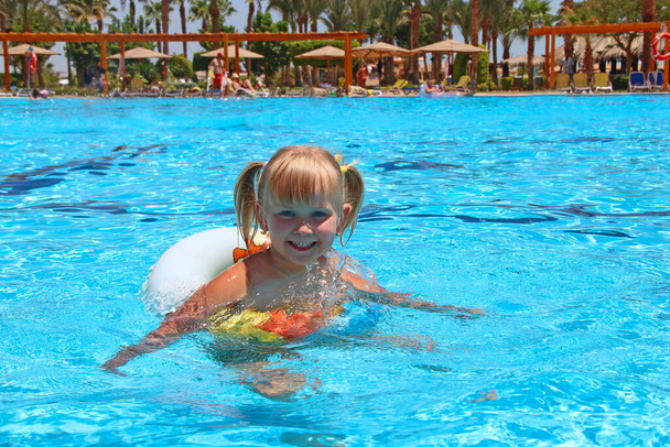 Улыбчивая девочка с приятной улыбкой плавает в бассейне, лежа на надувном круге на летних каникулах. Счастливого детства. Ребенок отдыхает на резиновом круге на голубой воде бассейна во время отдыха
 - Фото, изображение