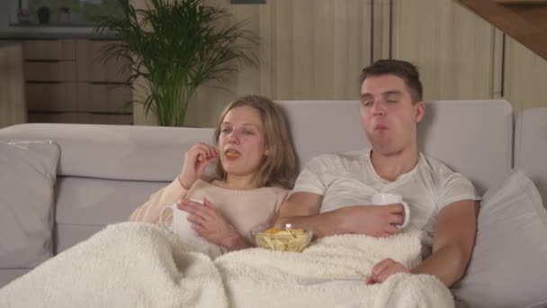 Tatlı bir çift abur cubur yiyor ve komik TV şovu izlerken çay içiyor. Genç çift kanepede oturuyor ve rahat bir sonbahar akşamında evinde film gecesinin tadını çıkarıyor.. - Video, Çekim