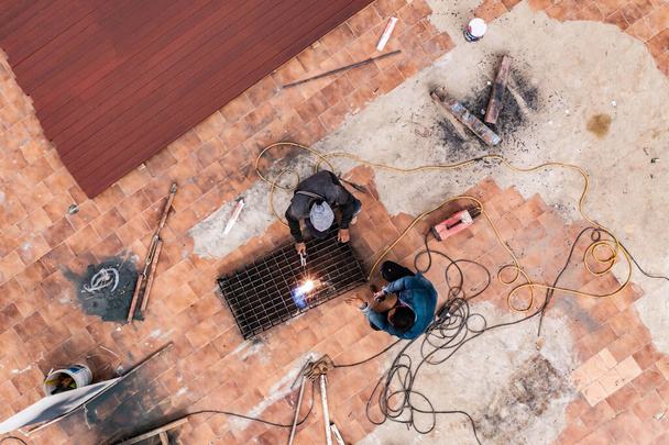 La foto aérea de dos hombres en uniforme gris y azul es metal de soldadura con máquina de soldadura por arco en el suelo en el sitio de trabajo. - Foto, imagen