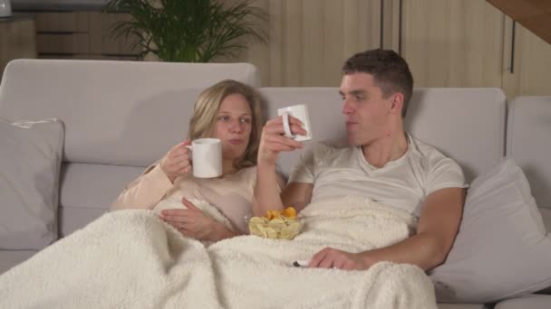 Κοντινό πλάνο: Νεαρό ζευγάρι χαλαρώνει σε έναν άνετο καναπέ και βλέποντας τηλεόραση ψυχαγωγίας. Twosome στον καναπέ και καλύπτονται με κουβέρτα απολαμβάνοντας σε χαλαρωτική ταινία νύχτα στο σπίτι σαλόνι σε ένα μακρύ βράδυ του χειμώνα. - Πλάνα, βίντεο