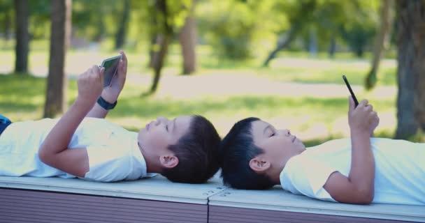 Kaksi poikaa leikkii älypuhelimilla ulkoilmakävelyn aikana. He makaavat penkillä kädet älypuhelimillaan. 4k kuvamateriaali. - Materiaali, video