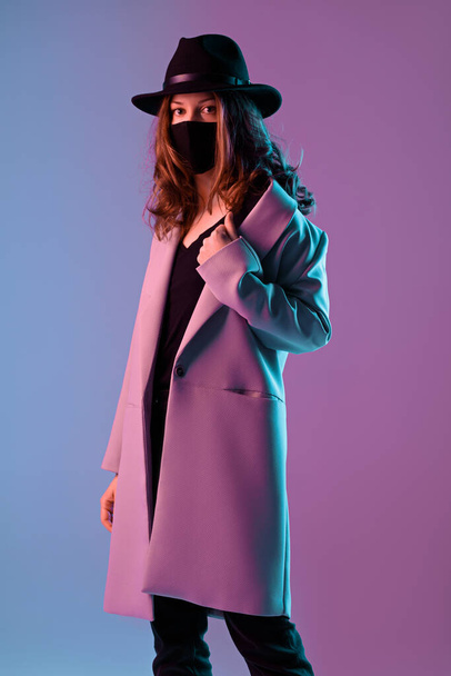 Portret van een trendy jonge vrouw in een jas, hoed en beschermend masker in paarse neon verlichting. - Foto, afbeelding