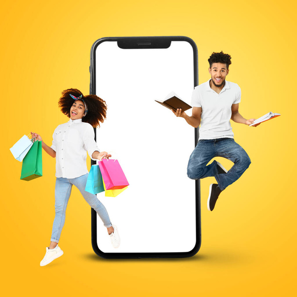 黄色の背景にショッピングバッグ、本、大きなスマートフォンでアフリカ系アメリカ人のカップルをジャンプ - 写真・画像