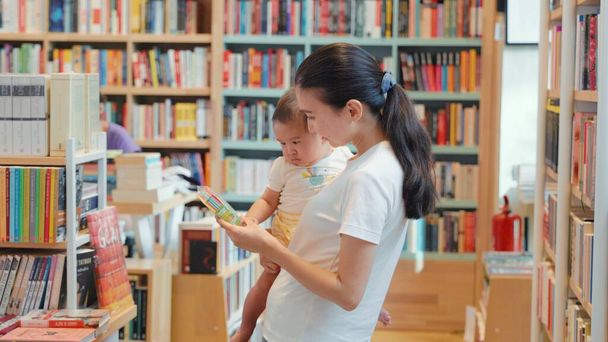 A fiatal anya egy színes könyvet mutat a babának egy könyvesboltban. Kiváló minőségű 4k felvételek - Fotó, kép
