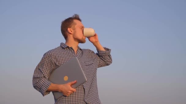 Macho caucasiano barbudo alegre em pé ao ar livre em pijama olhando para a frente, bebendo café ou chá segurando laptop. Conceito de manhã relaxante ou arrepiante. Imagens de vídeo 4k de alta qualidade - Filmagem, Vídeo