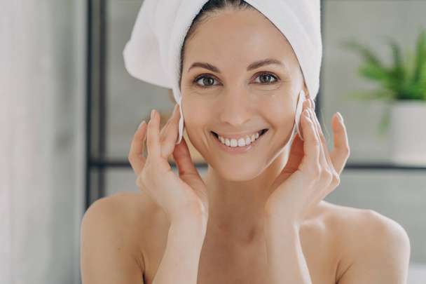 Pretty female with towel on head cleansing face, removing makeup, using soft cotton pads. Smiling hispanic woman wiping cheeks with moisturizing lotion or skin cleanser. Beauty treatment, skincare. - Photo, Image