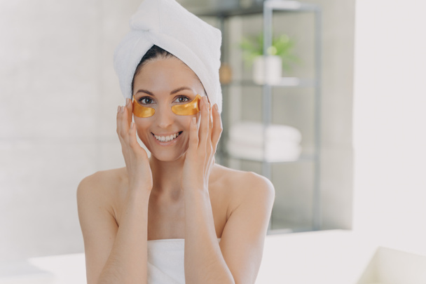 Hembra aplicando parches anti arrugas hidrogel dorado debajo de los ojos para suavizar la piel sana después de la ducha, haciendo rutina matutina cuidando la piel en el baño. Cuidado de la piel, concepto de tratamiento de belleza. - Foto, Imagen
