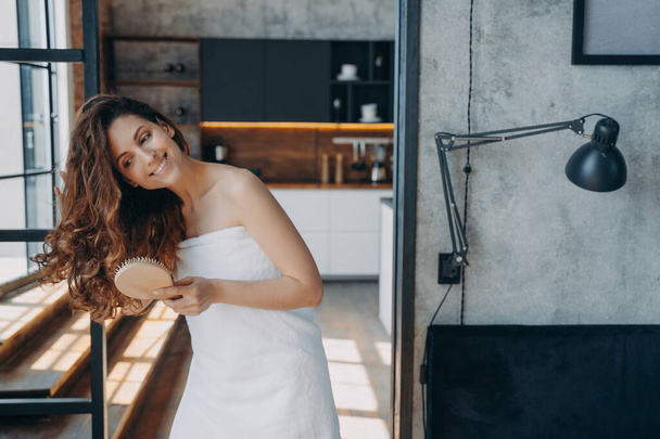 Smiling young female brushing healthy long brown hair with eco wooden brush. Happy latina woman wrapped in towel after shower getting ready for work, makes hairstyle at home. Haircare beauty treatment - Foto, imagen