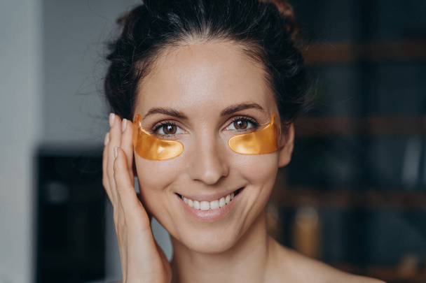 Mujer latina joven sonriente aplicando parches de colágeno hidratante dorados debajo de los ojos, feliz bonita hembra hace procedimiento de belleza facial anti arrugas en casa, cerca de retrato. Régimen de cuidado de la piel. - Foto, Imagen