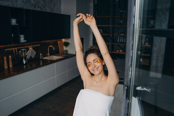 Entspannt lächelnde junge Frau verwendet unter den Augen Anti-Falten-Pflaster im Gesicht und dehnt sich in der Küche nach der Dusche. Glückliche Mädchen genießen morgendliche Beauty-Routine für gesunde Haut. Hautpflege.  - Foto, Bild
