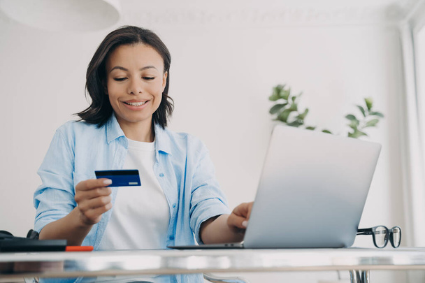 Női bankkártya mosolygás online banki szolgáltatásokkal a laptopon. Boldog nő fizet vásárol az interneten a számítógépen, ellenőrzi a pénzvisszafizetési bónuszt a munkahelyen. E-kereskedelem, ebusiness. - Fotó, kép