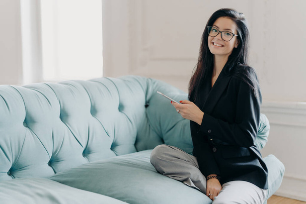 Happy business lady in official clothes, sits on comfortable sofa, uses mobile phone, reads news via network, searches information in cellular, going to phone partner, explores online project - Photo, Image
