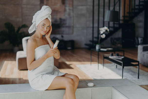 Pleased healthy woman applies body lotion and face cream, nourishes skin, wrapped in bath towel, smiles gently, poses against domestic interior, undergoes beauty treatments. Dermatology concept - Photo, Image