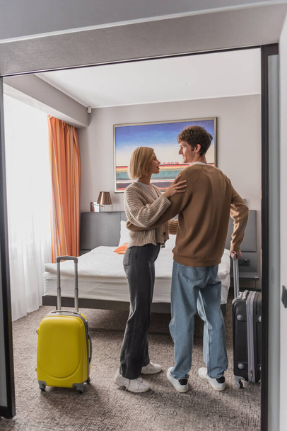 full length of happy young couple looking at each other near baggage in hotel bedroom - Photo, image
