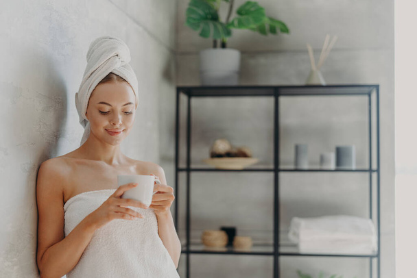 Fresh young European woman wrapped in towel after bath, stands in bathroom with mug of coffee or tea, enjoys morning, has pleased face expression, healthy glowing skin. Hygiene routine concept - Photo, Image