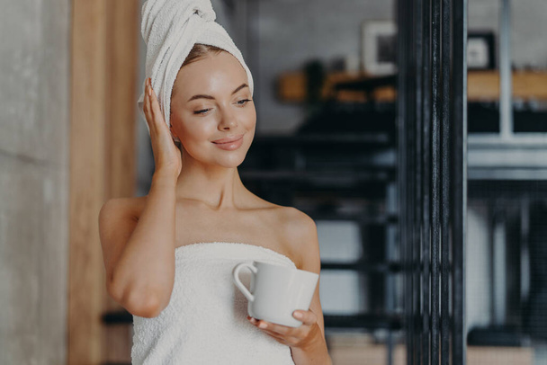 Φωτογραφία της υγιούς στοχαστική γυναίκα με υγιή λείο δέρμα ποτά ζεστό τσάι συμπυκνωμένο κάτω, στέκεται τυλιγμένο σε λευκή πετσέτα μπάνιου στο άνετο δωμάτιο, απολαμβάνει την υγιεινή και την ομορφιά θεραπείες, αισθάνεται χαλαρή - Φωτογραφία, εικόνα