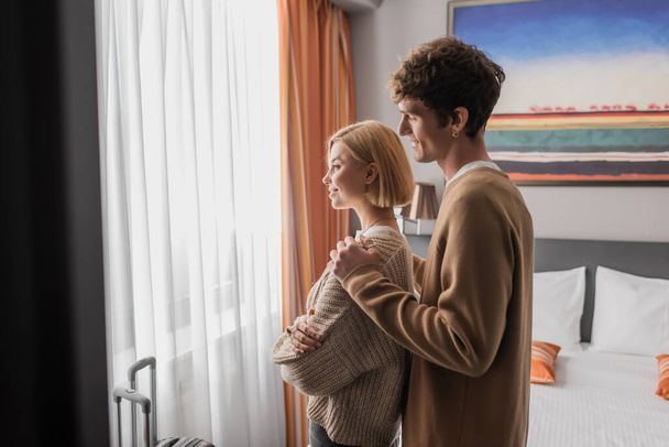 side view of smiling man hugging girlfriend standing with crossed arms near window in hotel room - Foto, Imagen
