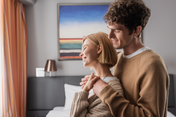 side view of brunette man embracing young girlfriend in hotel bedroom - Foto, immagini