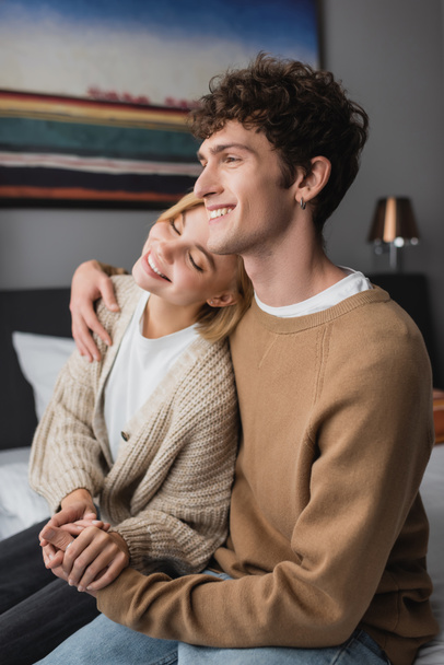 Улыбающийся мужчина, держащийся за руки со счастливой девушкой, сидящей с закрытыми глазами на кровати в отеле - Фото, изображение