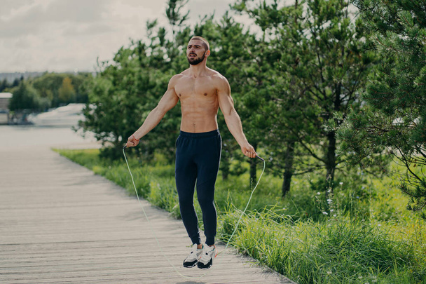 Sportowy mężczyzna z grubą brodą wykonuje ćwiczenia ze skakanką, ma muskularny tułów, pozostaje w dobrej kondycji fizycznej, stwarza na świeżym powietrzu. Zdrowa i sprawna koncepcja. mężczyzna biegacz rozgrzewa się z w sprzęt sportowy - Zdjęcie, obraz