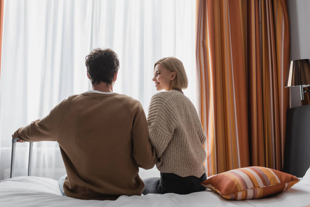 back view of happy woman looking at brunette boyfriend while sitting in hotel bedroom - Fotoğraf, Görsel