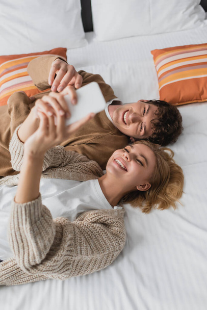 вид на молодую веселую пару, делающую селфи на размытом мобильном телефоне, лежащую на кровати в отеле - Фото, изображение