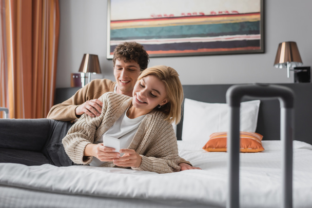 smiling woman using mobile phone near happy young boyfriend on bed in hotel apartments - Foto, Imagem