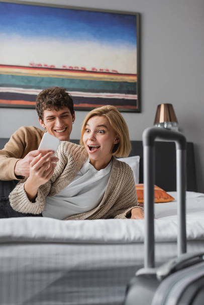 smiling man showing mobile phone to amazed girlfriend on bed in hotel apartments - Fotografie, Obrázek