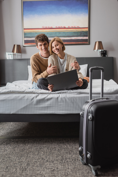 cheerful couple of travelers looking at camera while sitting on bed with laptop near suitcase in hotel room - Foto, Imagem