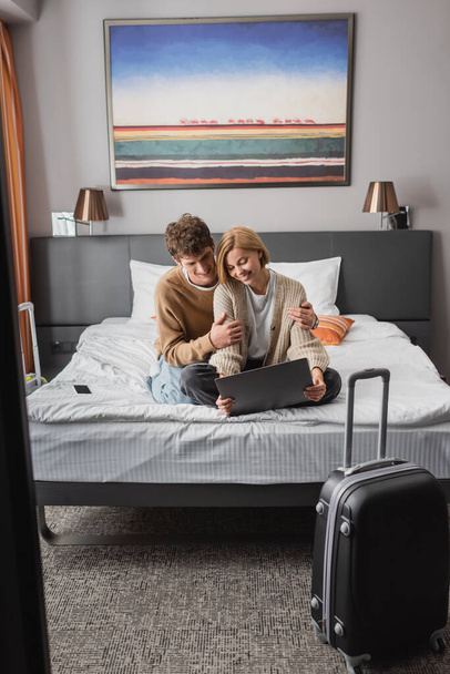happy woman sitting with laptop near smiling boyfriend on bed in modern hotel - Foto, afbeelding