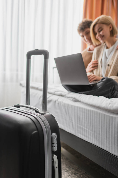 selective focus of travel bag near blurred couple watching movie on laptop in hotel bedroom - Photo, image