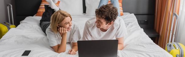 cheerful young couple looking at each other near laptop and smartphone on bed in hotel suite, banner - Fotó, kép