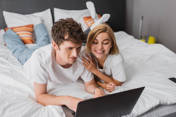 молодая радостная пара смотрит фильм на ноутбуке, лежа на кровати в апартаментах отеля - Фото, изображение