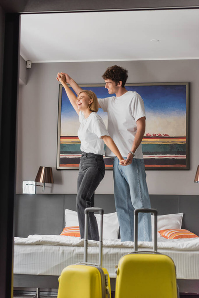 young and cheerful couple holding hands and having fun while standing on bed in modern hotel apartments - Foto, afbeelding