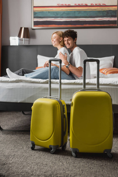 selective focus of travel bags near happy young couple sitting on hotel bed on blurred background - Фото, изображение