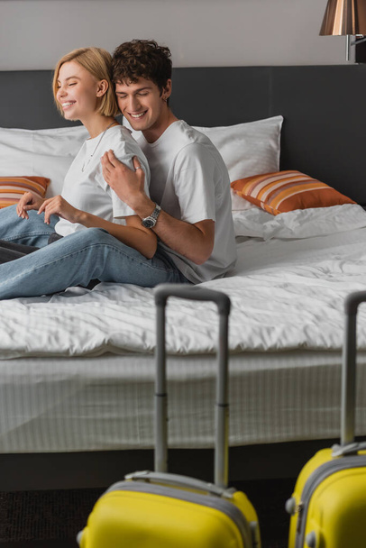 pleased man embracing girlfriend while sitting on hotel bed near suitcases on blurred foreground - Photo, Image