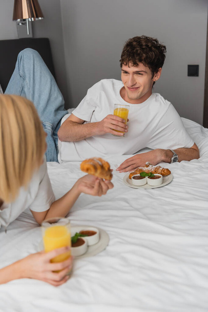 cheerful man holding orange juice and looking at blonde girlfriend having breakfast on hotel bed - Photo, Image